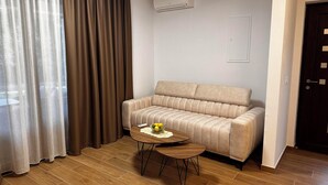 A room with a beige sofa, a small round wooden table with a bowl of fruit, a window with sheer curtains, and a dark door. - Apartments Robi 2 (Rovinj)