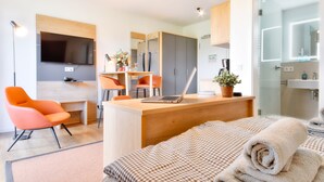 A modern hotel room with a bed, a desk with a laptop, a TV, and a bathroom with a mirror and sink.