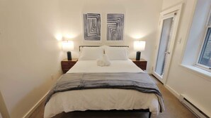 A neatly made bed with a white and gray blanket, two bedside lamps, and abstract wall art. - Capitol Hill & Penn - Steps from Eastern Market (Washington)