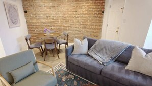 A living room with a brick wall, a blue sofa, a wooden chair, a round table with chairs, and a patterned rug. - Capitol Hill & Penn - Steps from Eastern Market (Washington)
