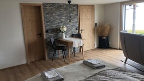 Et soveværelse med seng, et natbord, en stol, et lille bord med blomster i en vase, og et vindue med udsigt udendørs.