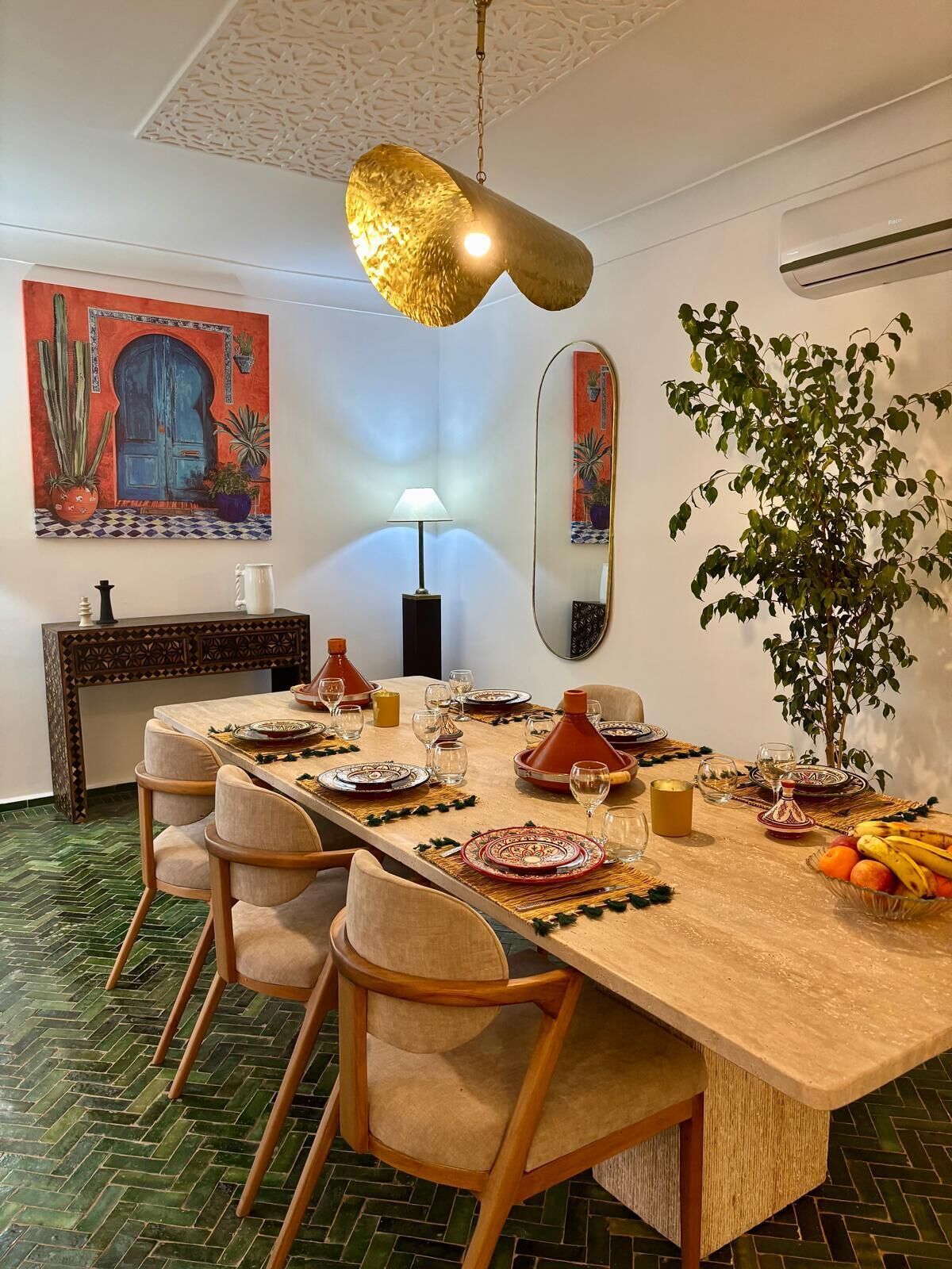 A dining area with a long table set for a meal, surrounded by chairs, a hanging light fixture, and decorative elements including a mirror and artwork.