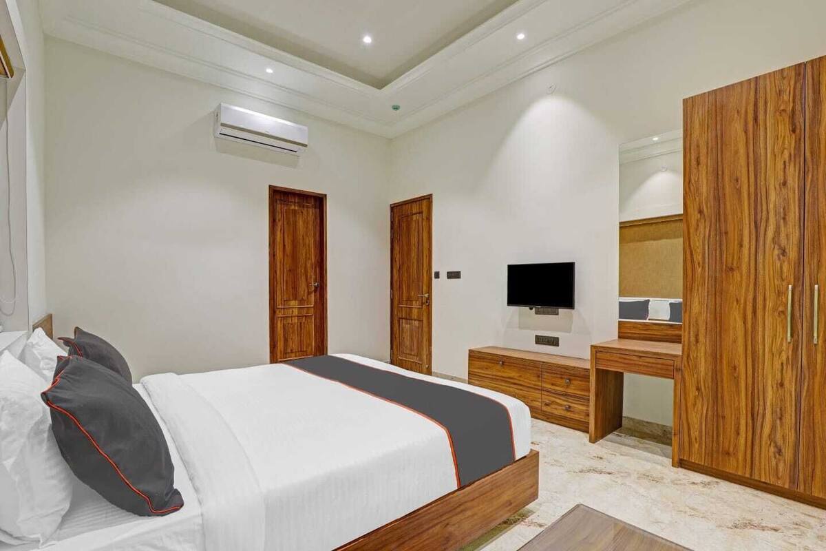 A bedroom with a bed, wooden wardrobe, a desk with a flat-screen TV, and an air conditioning unit.