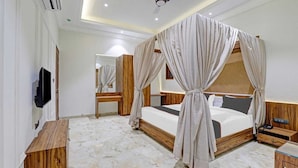 A bedroom with a four-poster bed, wooden headboard, a flat-screen TV mounted on the wall, a wooden nightstand, and a marble floor. - HOTEL RATNA VILAS PALACE (Rajkot)