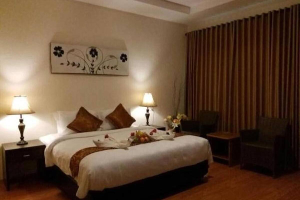 Quarto de hotel com cama grande, duas luminárias laterais, um objeto decorativo na parede, uma cadeira e cortina.