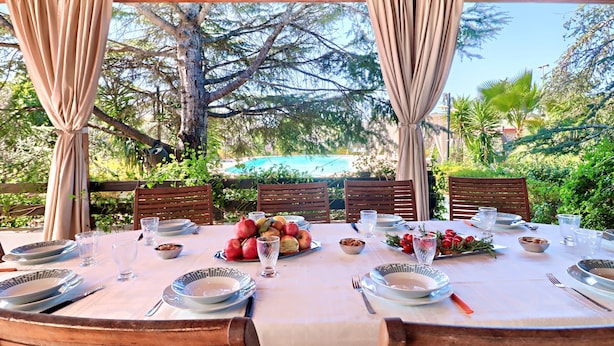 A table set for dining with plates, glasses, and a fruit bowl, surrounded by wooden chairs and curtains, with a view of trees and greenery. - Villa Velisia - Con Ampia Piscina E Campo Da Gioco Privato (Alessano)