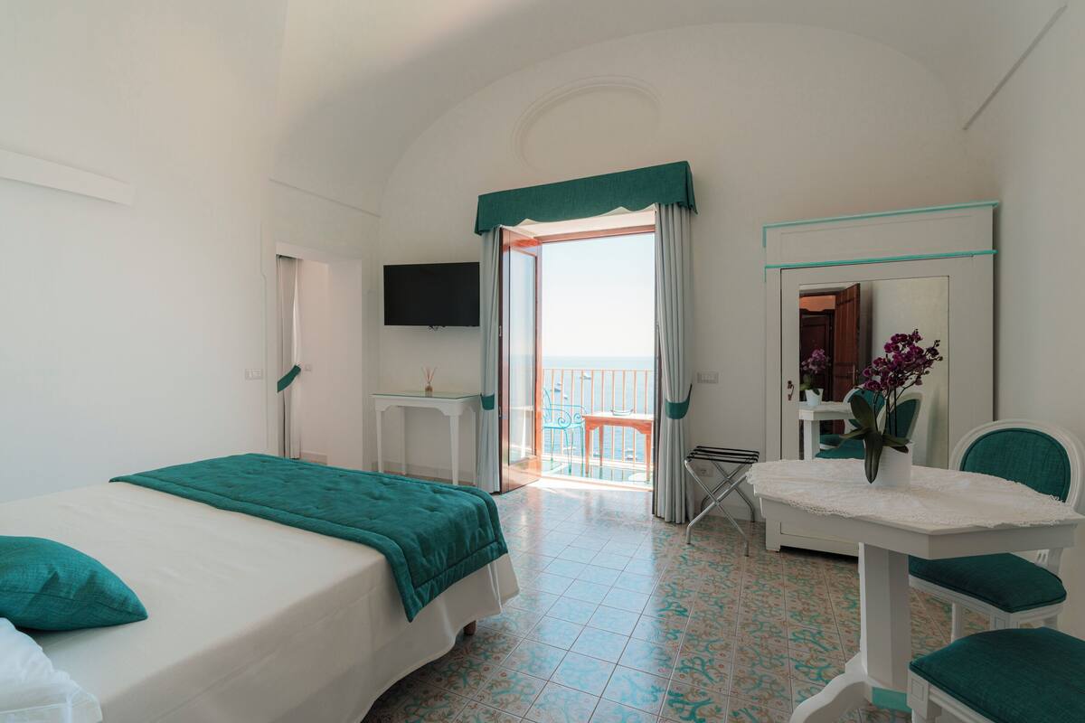 A hotel room with a balcony, a bed, a desk, a chair, and a mirror.