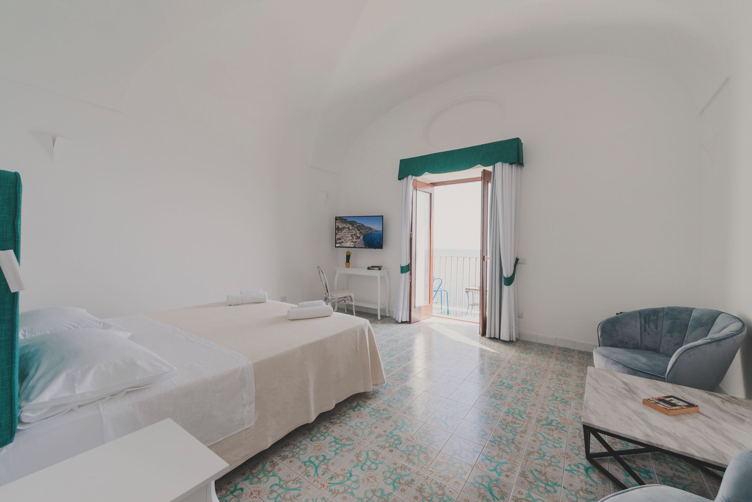 Quarto com cama, cadeira, mesa pequena e vista para o mar através de uma porta aberta.