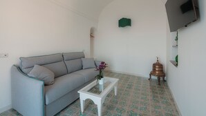 A living room with a grey sofa, a white coffee table with a vase of flowers, patterned floor tiles, a flat-screen TV mounted on the wall, and a small decorative item on the floor. - Villa Nettuno (Positano)