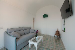 Una sala de estar con un sofá gris, una mesa de centro blanca con un jarrón de flores, baldosas de piso estampadas, un televisor de pantalla plana montado en la pared y un pequeño objeto decorativo en el suelo.