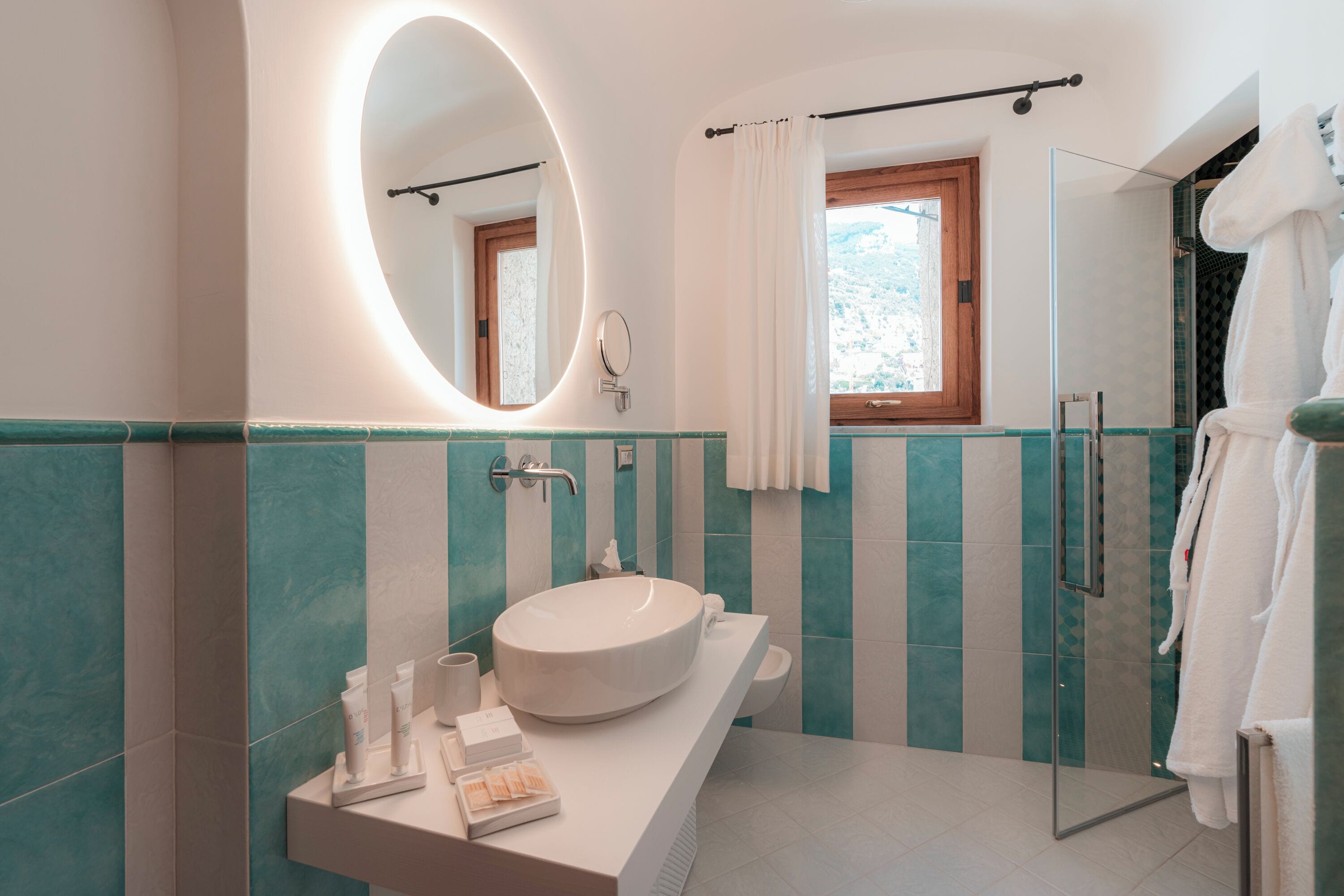Une salle de bain moderne avec un grand lavabo ovale, un miroir rond, une cabine de douche en verre et une fenêtre avec un rideau.