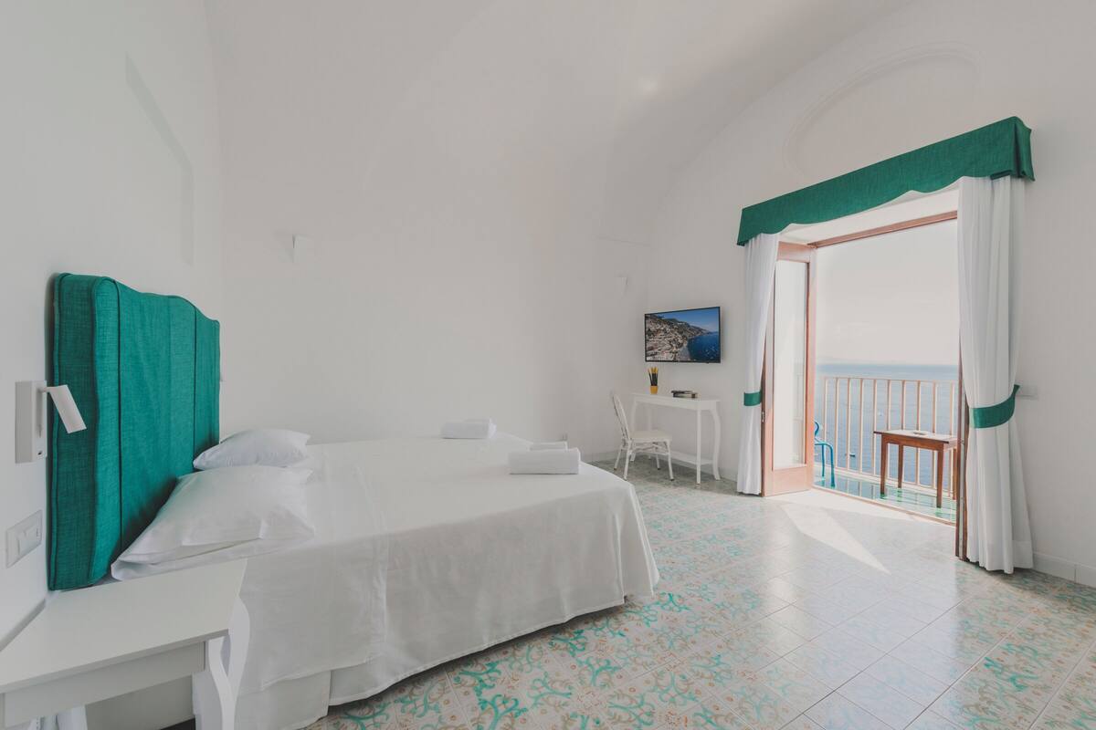 A hotel room with a bed, a desk, a chair, and a balcony with a view of the sea.