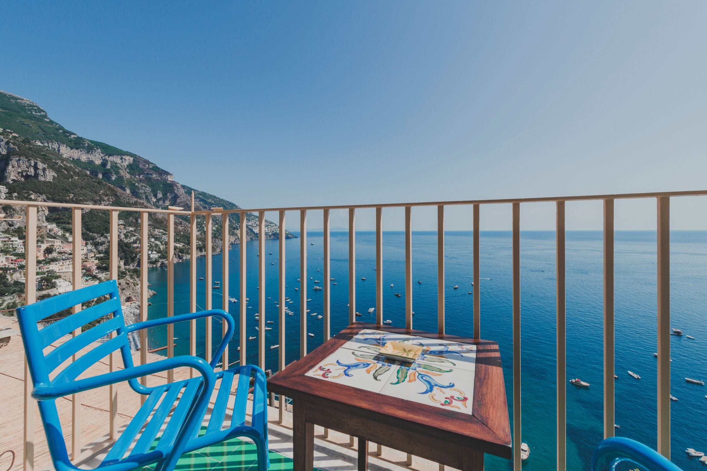A balcony with a view of the sea, a table with a tile top, and blue chairs.