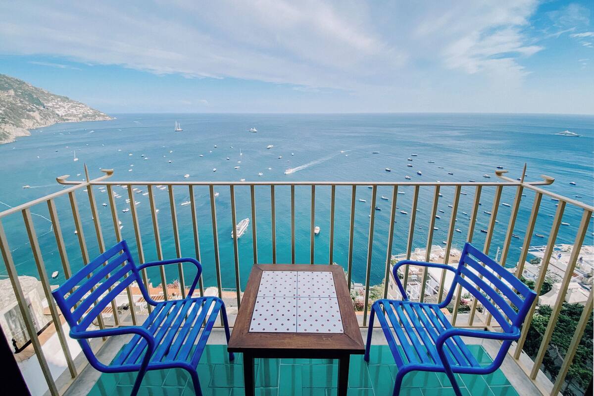 A balcony with two blue chairs and a table, overlooking a coastal view with boats and a clear sky.