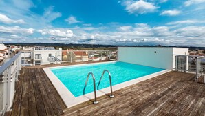 Une piscine sur le toit, avec une terrasse en bois, des rampes en métal et un ciel d’un bleu limpide.