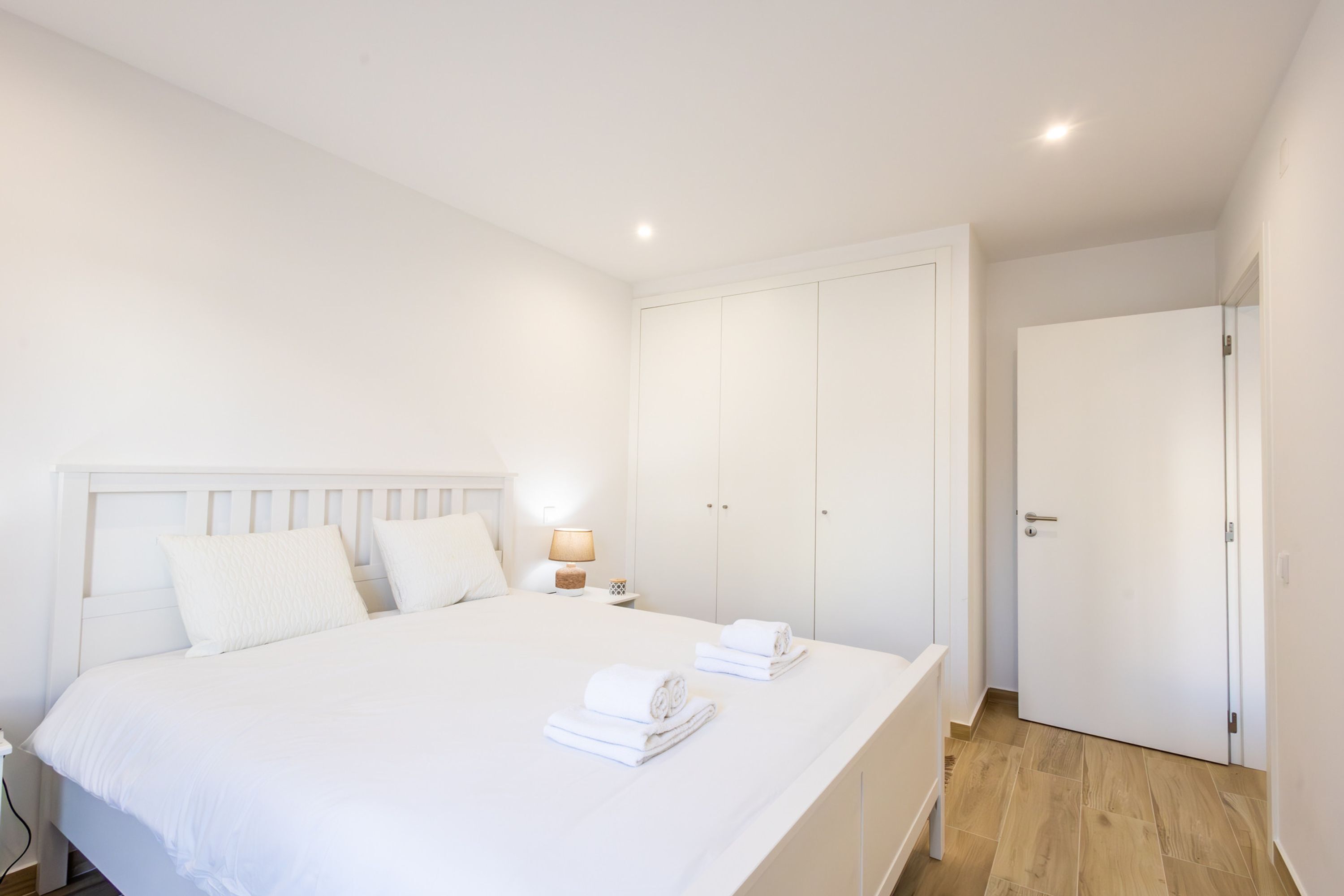 Un dormitorio con una cama grande, cabecera blanca, dos mesitas de noche con lámparas, un armario y una puerta.