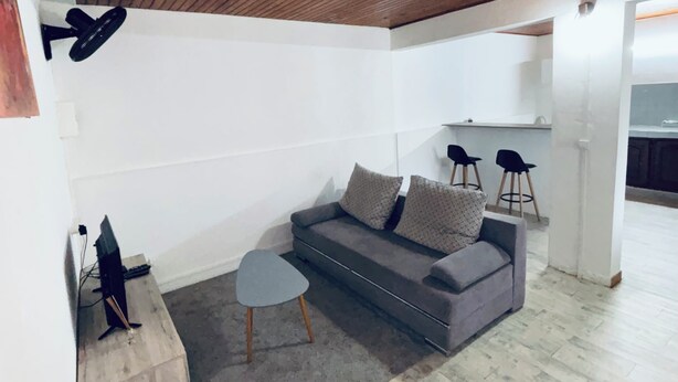 A modern living room with a wooden ceiling, a grey sofa, a small round table, and two black chairs. - Comfortable air-conditioned mahogany T2 Lamentin near Galleria shopping center (Le Lamentin)