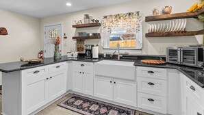 Una cocina moderna con armarios blancos, encimeras negras y un horno empotrado. Hay repisas sobre el mostrador con varios utensilios de cocina, y una ventana con un cortinaje decorativo.