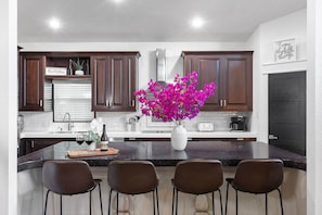 Une cuisine avec des meubles en bois foncé, un plan de travail blanc, un îlot central avec quatre chaises, un vase rempli de fleurs roses et une machine à café.