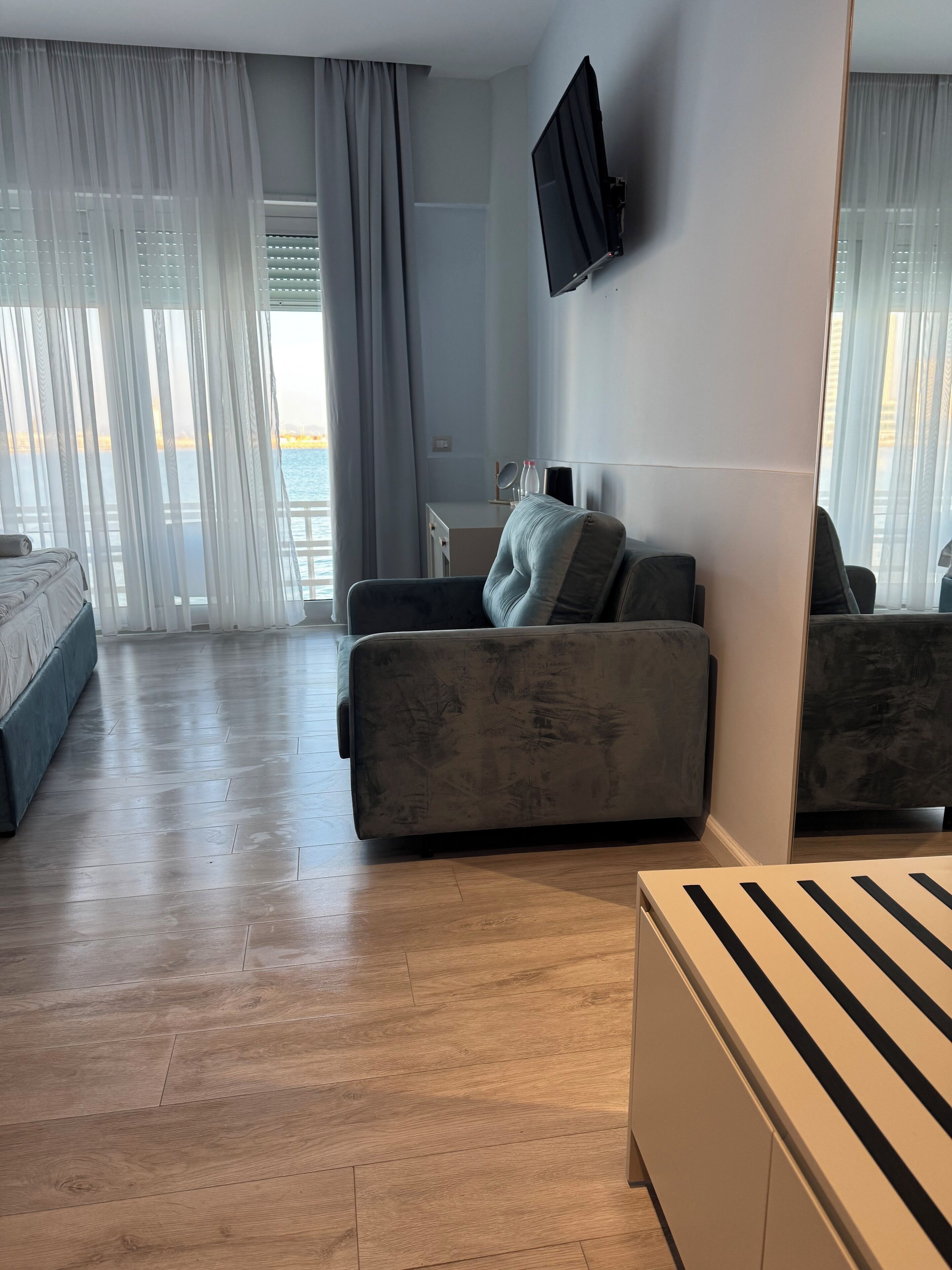 A modern hotel room with a sofa, a bed, a flat-screen TV, and a large window with curtains.