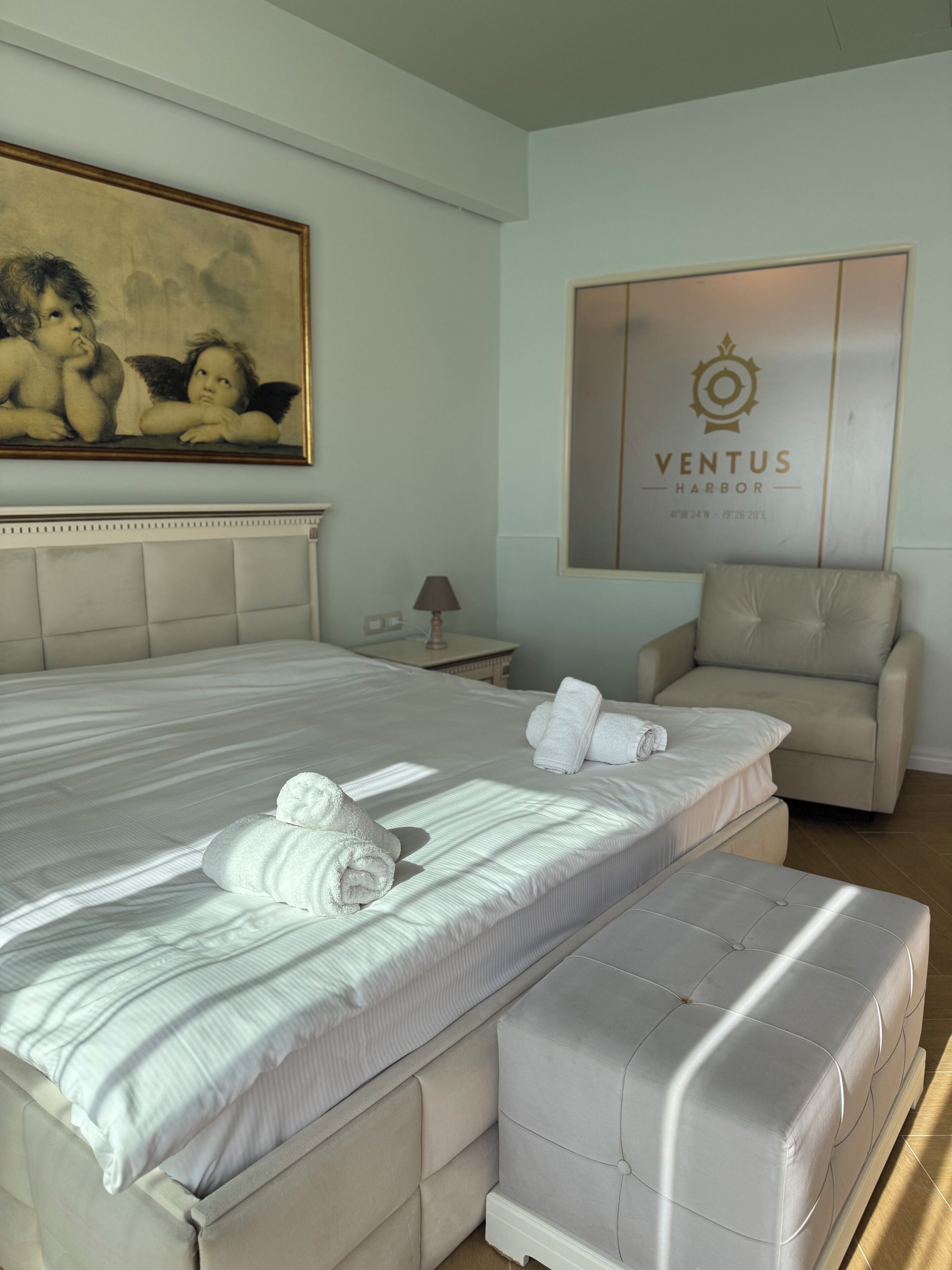 A hotel room with a large bed, a chair, a painting, and a wall with a logo.