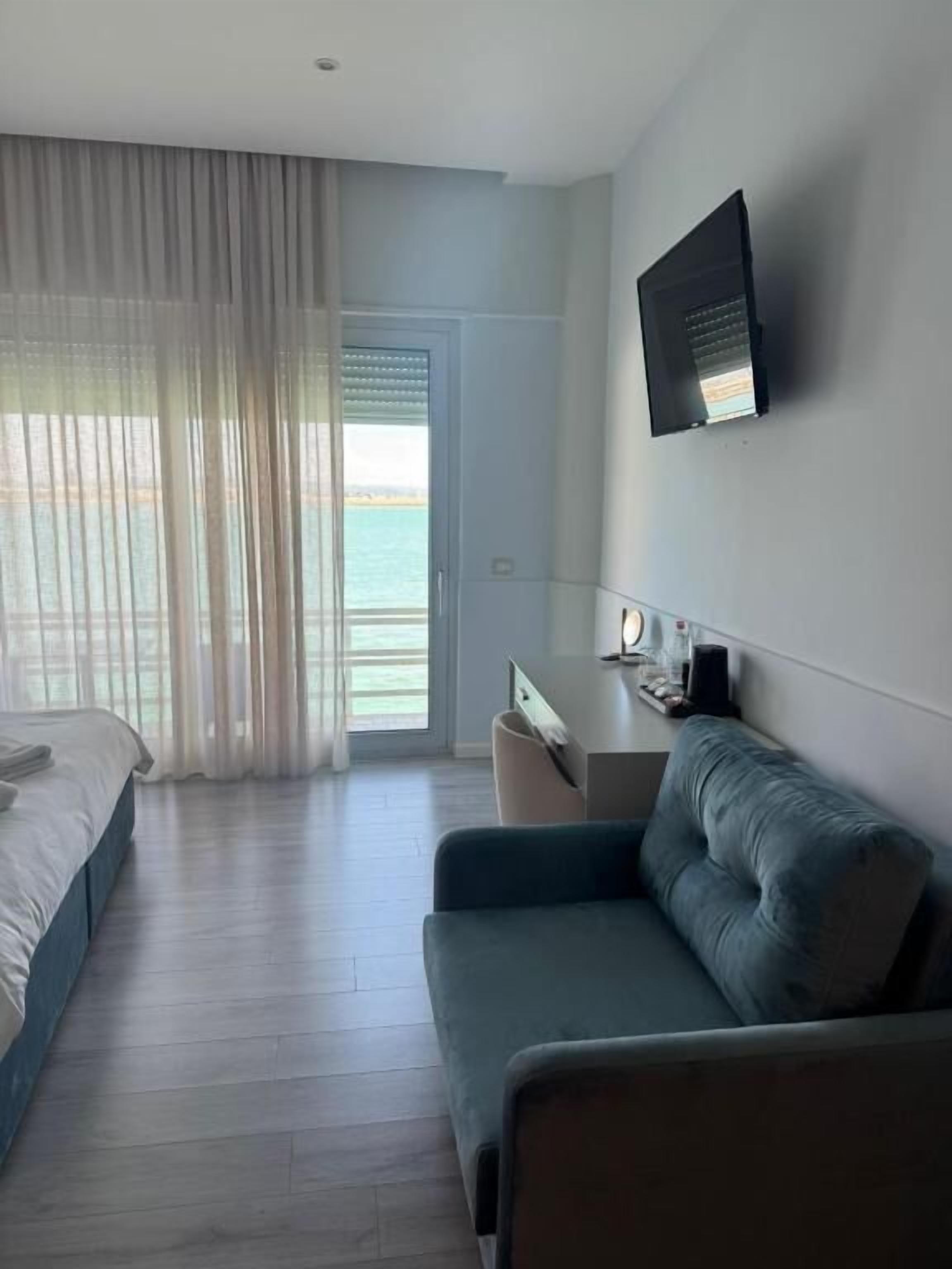 Habitación de hotel con cama, sofá, escritorio y vistas al mar a través de ventanas amplias.