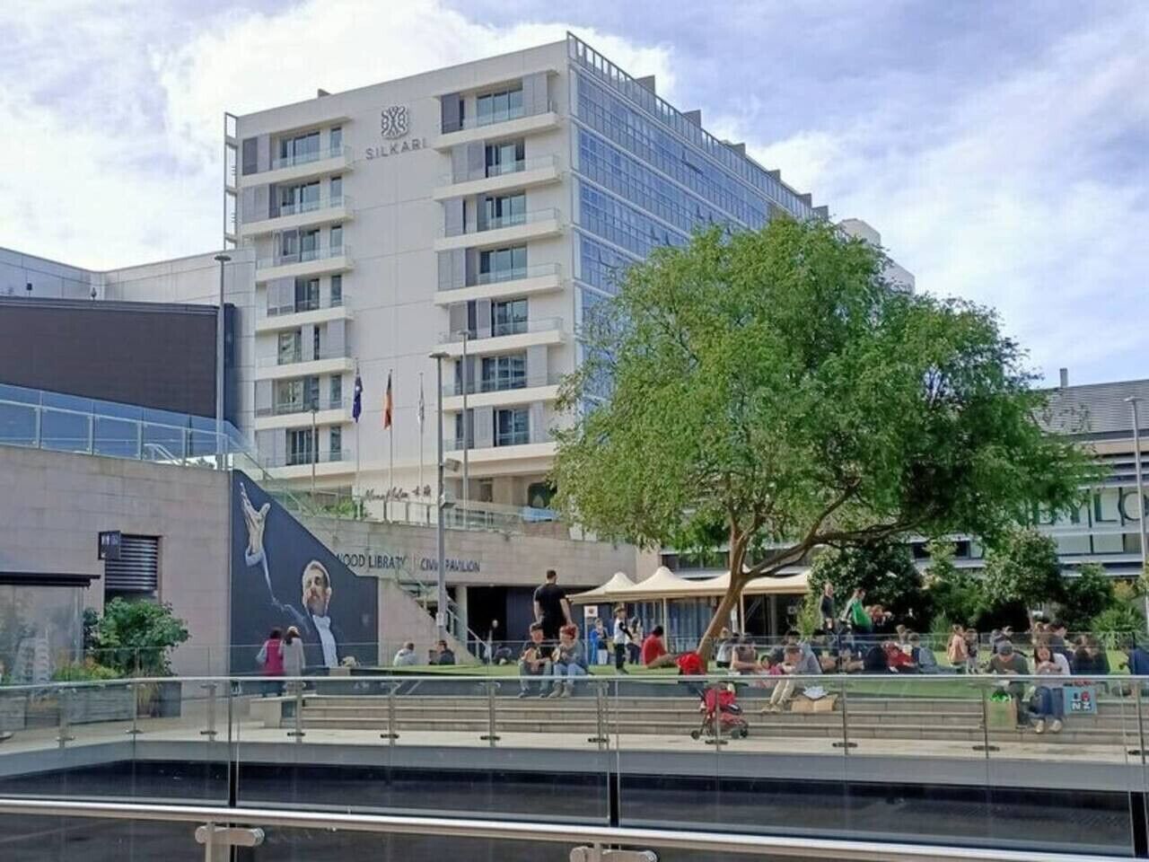 A modern building with multiple floors, a large tree in front, and people sitting and walking around.