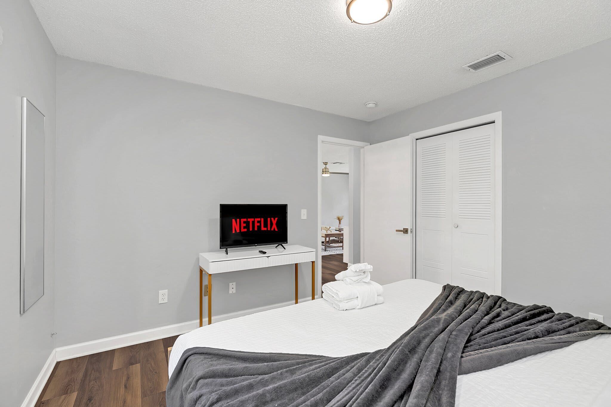 A bedroom with a bed, a TV displaying 'NETFLIX', a white bedside table, and a door leading to another room.