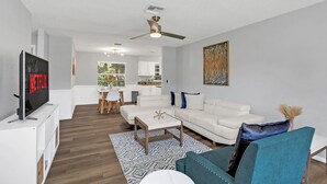A modern living room with a white sofa, a teal armchair, and a coffee table. A flat-screen TV is mounted on a white cabinet. The room has a ceiling fan, a painting on the wall, and a view into the kitchen area. - Backyard BBQ home - Blue Spring to Daytona (Orange City)