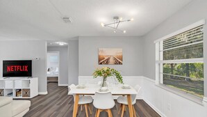 A modern living room with a flat-screen TV, a dining area with a white table and chairs, and a large window with blinds. - Backyard BBQ home - Blue Spring to Daytona (Orange City)