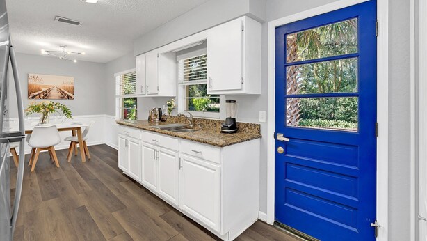 A modern kitchen with white cabinets, a granite countertop, and a blue door with a view of greenery. - Backyard BBQ home - Blue Spring to Daytona (Orange City)