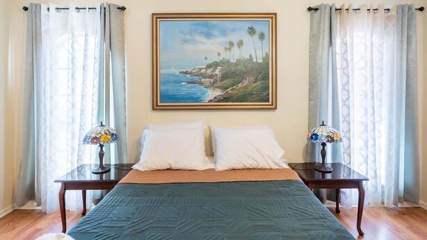 A bedroom with a bed, two nightstands, a painting of a coastal landscape, and sheer curtains. - Spacious 4BR with Private Pool, Near Six Flags (San Antonio)