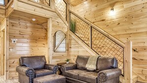 L’intérieur chaleureux d’un chalet en bois présente un grand canapé en cuir, un fauteuil assorti et un pouf. Un escalier en bois avec une rampe en métal mène à l’étage supérieur.
