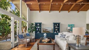 A spacious living room with a glass door leading to a garden, wooden ceiling beams, a white sofa, a wicker chair, and two blue shelving units. - Classic Cottage in the Heart of La Jolla Shores (San Diego)