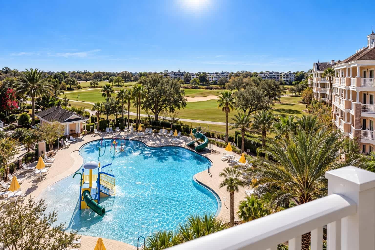 A swimming pool with a slide, surrounded by palm trees and lounge chairs, with a golf course in the background.