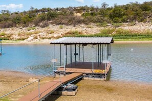 A dock with a covered seating area extending into a body of water, surrounded by a natural landscape. - Lake House | Pool, Hot Tub, Game Room, Scenic View (Spicewood)