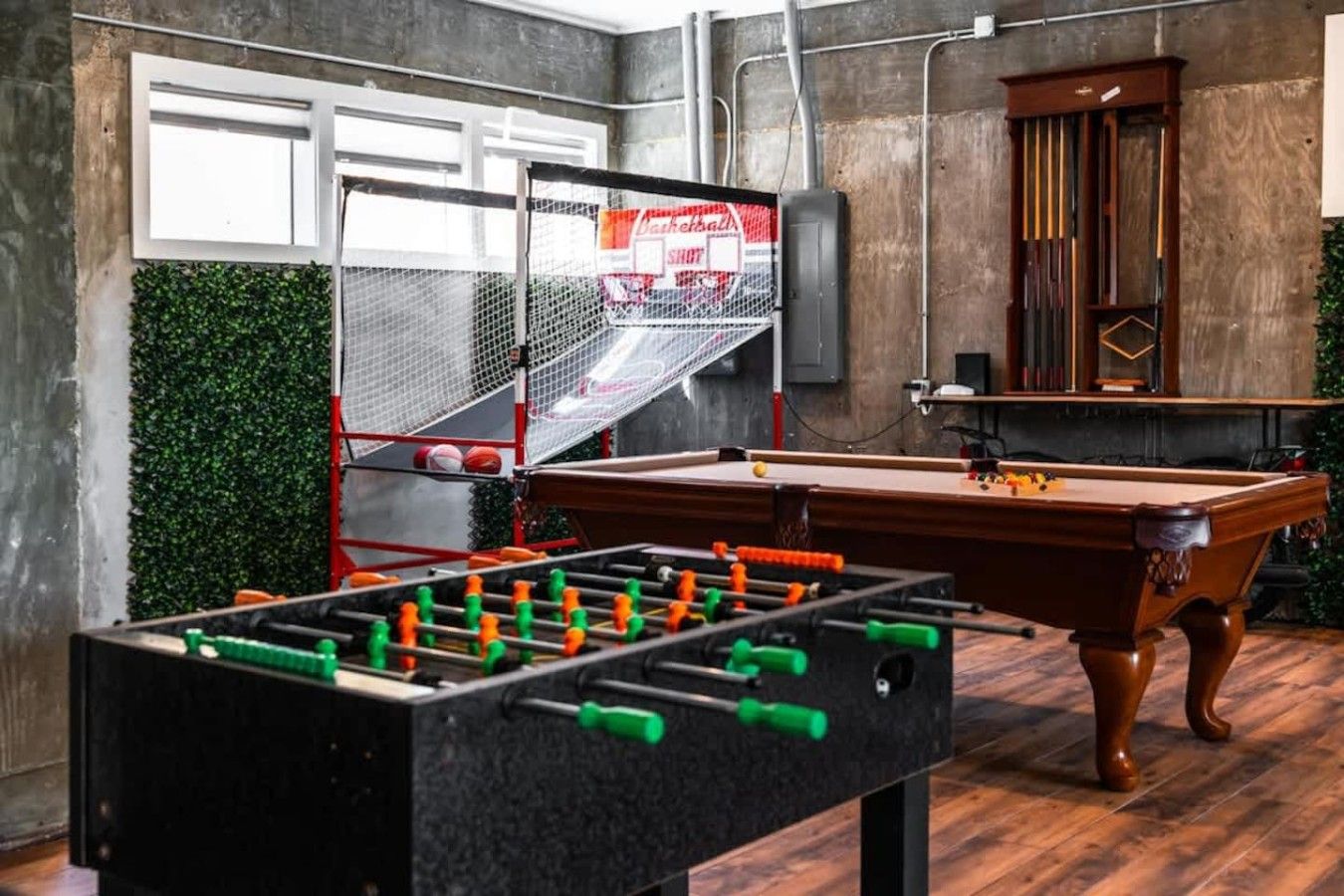 Mesa de bilhar, mesa de futebol de mesa e uma cesta de basquete em um espaço com parede de concreto e decoração verde na parede.