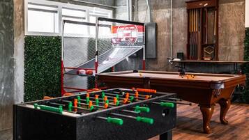 Mesa de bilhar, mesa de futebol de mesa e uma cesta de basquete em um espaço com parede de concreto e decoração verde na parede.