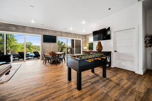 A spacious room with a foosball table, a dining area with chairs, and a television mounted on the wall. - Lake House | Pool, Hot Tub, Game Room, Scenic View (Spicewood)