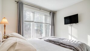 Un dormitorio con una cama, una lámpara, una ventana con persianas y un televisor montado en la pared.