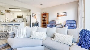 Una sala moderna con un sofá seccional, una estantería de madera, un sillón azul y una cocina al fondo.