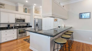 A modern kitchen with white cabinetry, stainless steel appliances, a large island with bar stools, and a view of the outdoors. - Gorgeous home with pool and golf cart included! Pet friendly! (Destin)