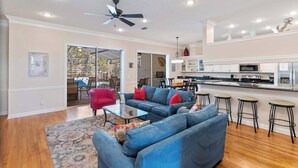 A spacious living room with a blue sofa set, a red armchair, and a glass coffee table. A kitchen with white cabinets and a bar area is visible in the background. - Gorgeous home with pool and golf cart included! Pet friendly! (Destin)
