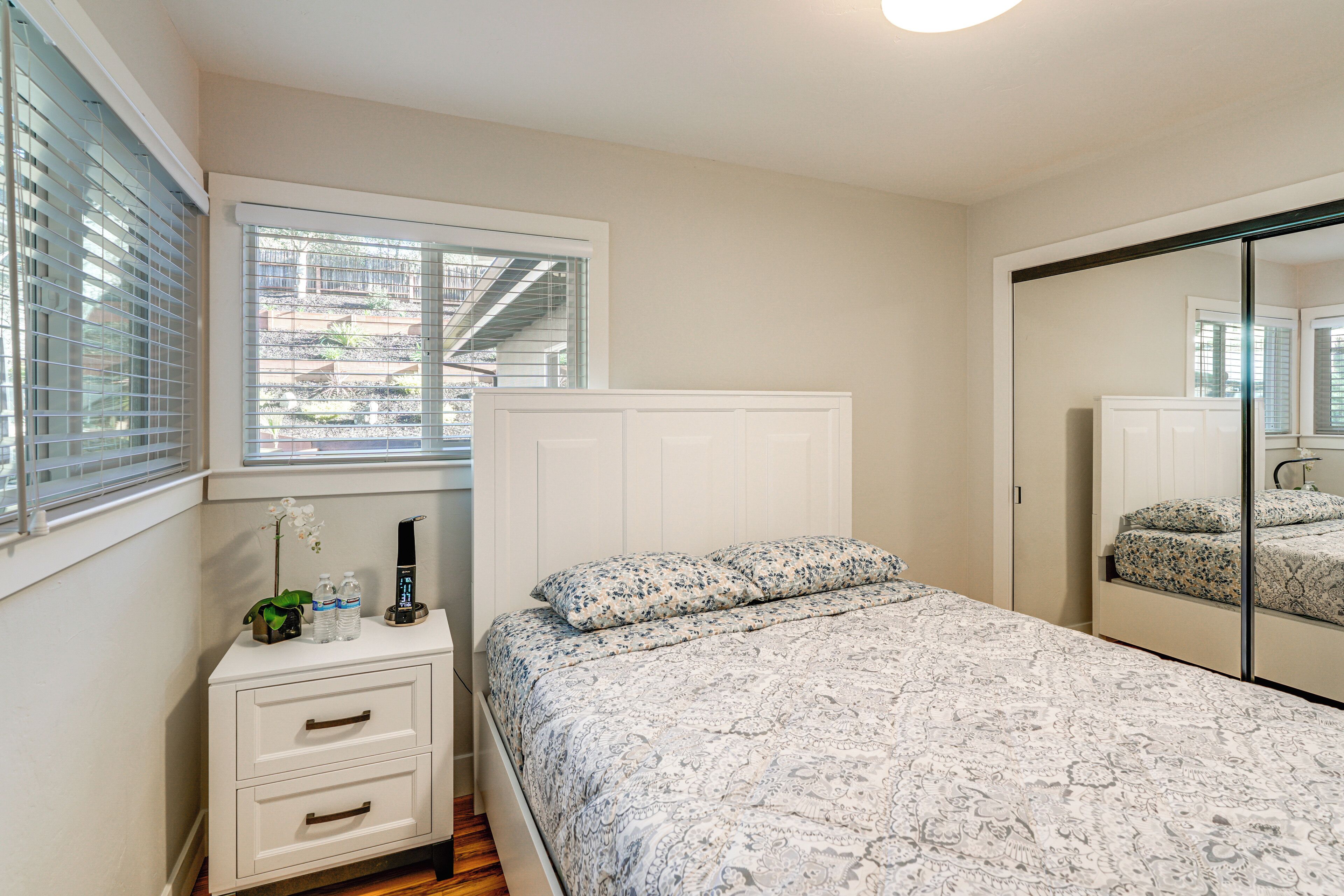 A bedroom with a bed, a bedside table, a mirror, and a window with blinds.