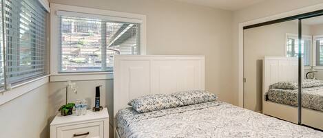 A bedroom with a bed, a bedside table, a mirror, and a window with blinds.