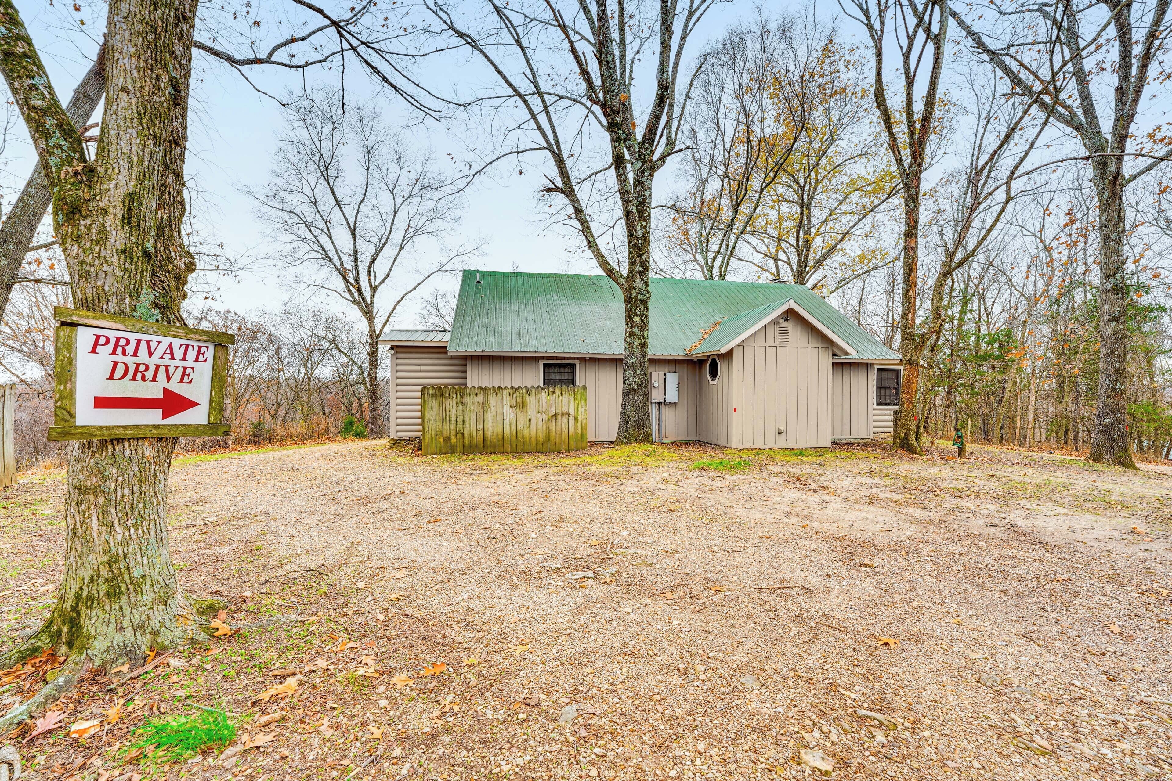 A private drive leading to a single-story house with a green roof, surrounded by trees and a gravel driveway.