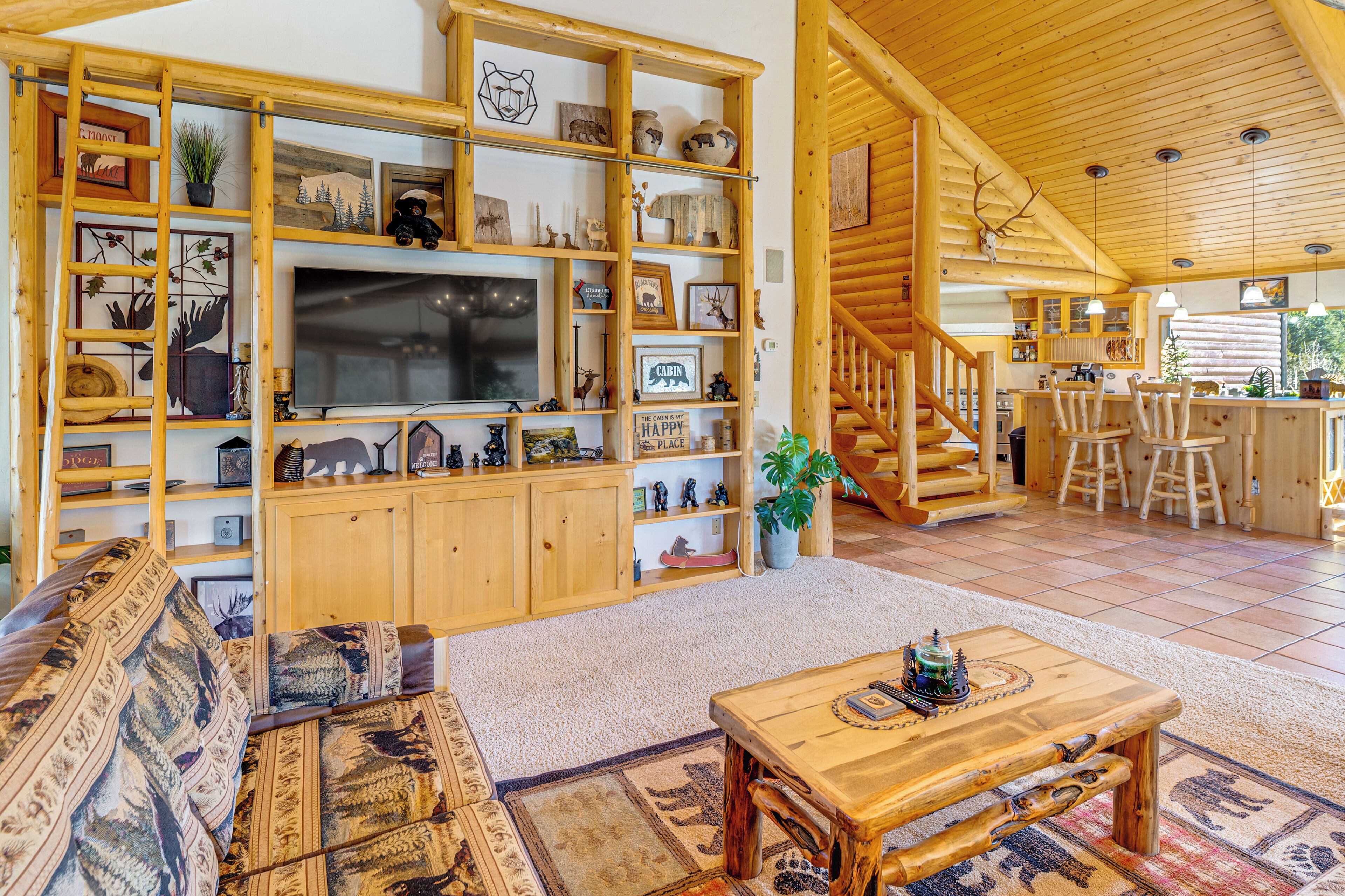 Un acogedor interior de cabaña con una escalera de madera, una zona de comedor y una sala de estar con un televisor grande y estanterías de madera llenas de objetos varios.