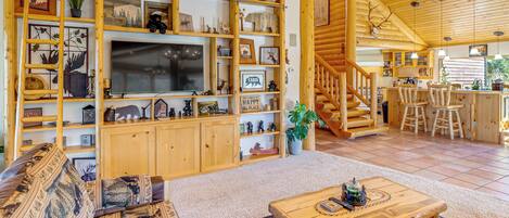 Un acogedor interior de cabaña con una escalera de madera, una zona de comedor y una sala de estar con un televisor grande y estanterías de madera llenas de objetos varios.