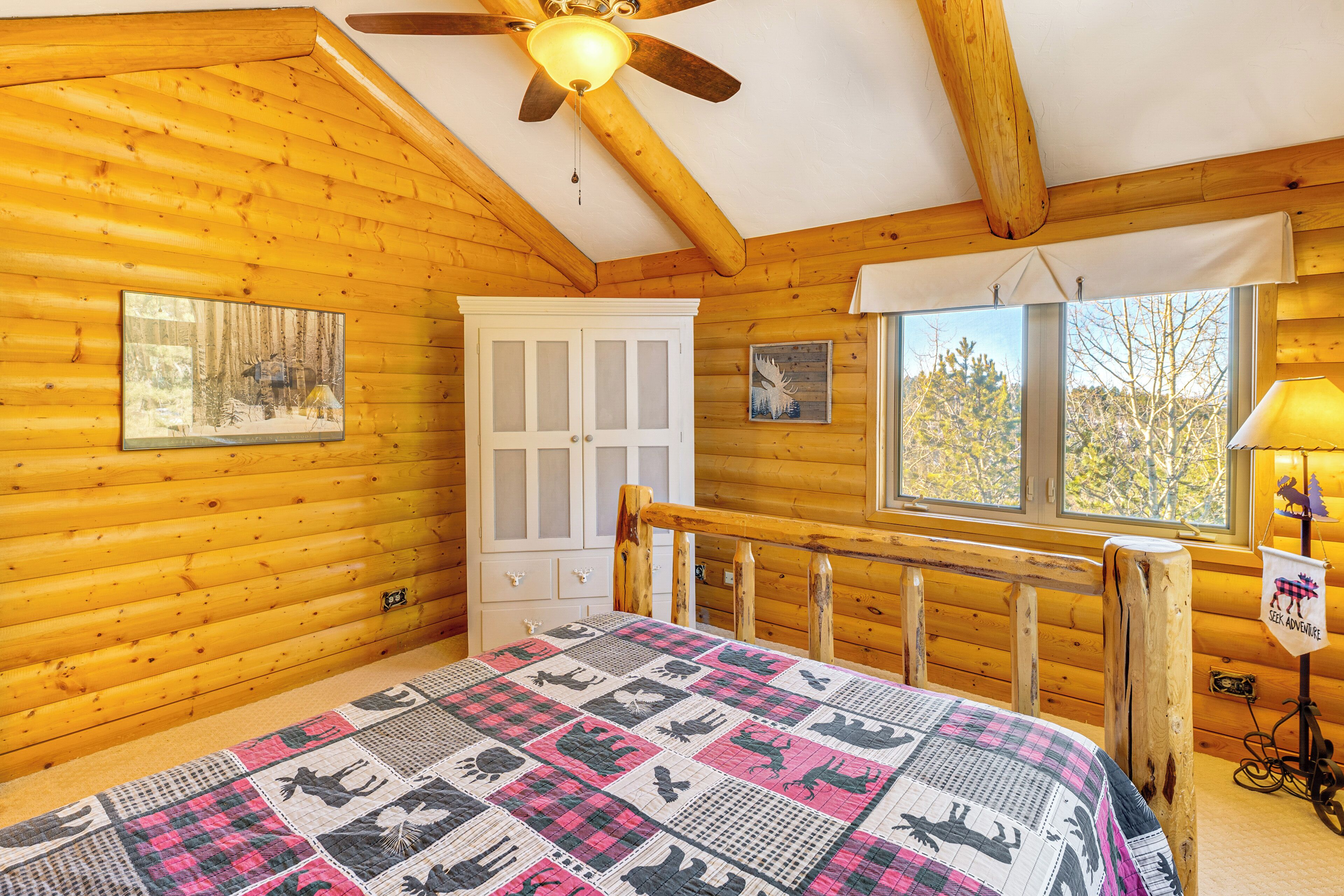 Une chambre située dans une cabane en rondins, avec un lit en bois, un ventilateur de plafond, une fenêtre donnant sur la forêt et une porte blanche.
