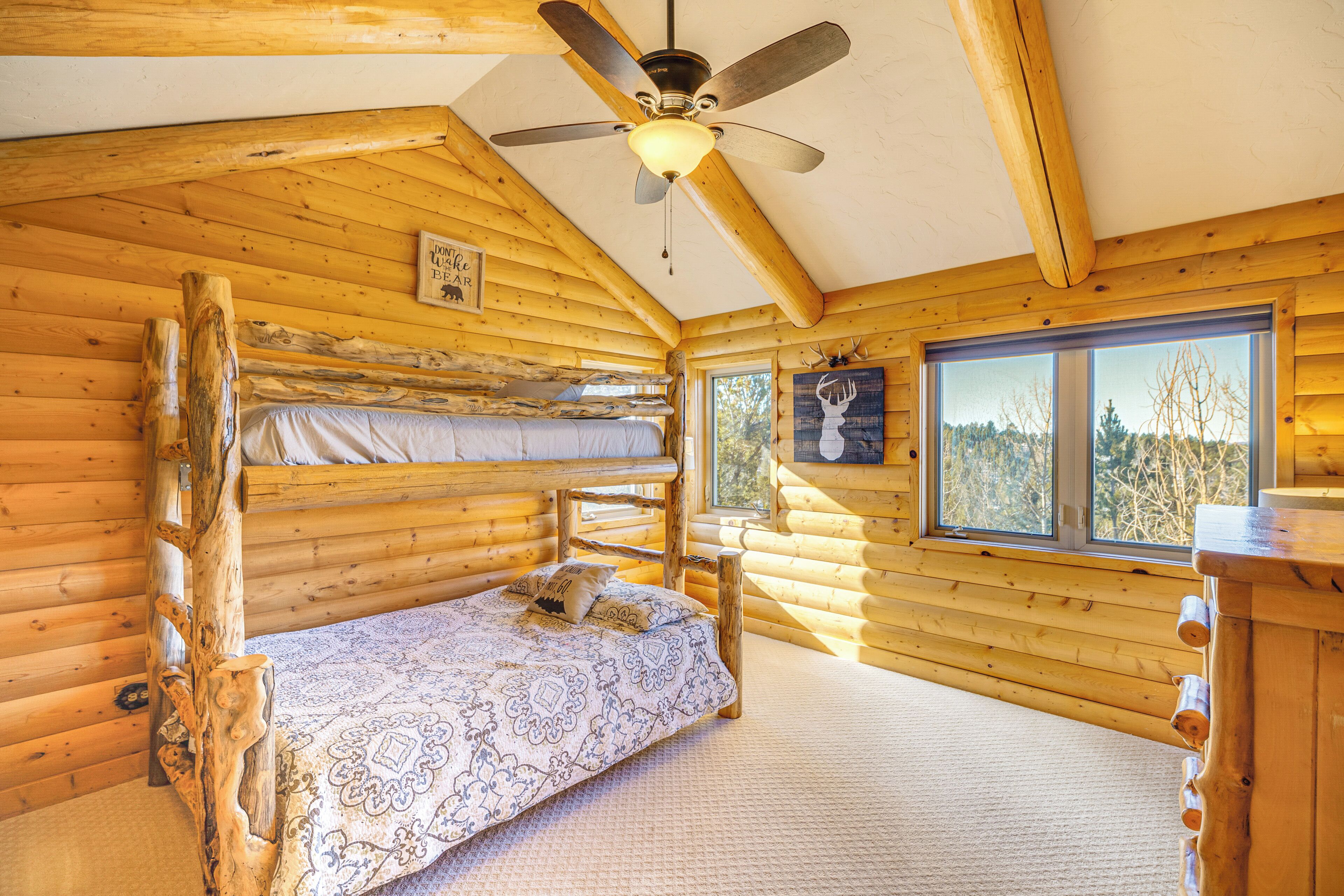 Quarto de cabana de tronco com uma cama beliche, um ventilador de teto e uma janela ampla com vista para árvores.